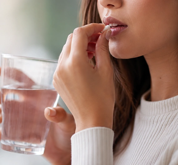 A close-up of a woman taking a white tablet with water, wearing a white ribbed sweater, with a glass of water in the background.